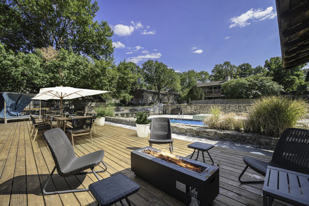 A wooden patio deck with a fireplace and chairs surrounded by greenery and a pool at Corinth Communities.